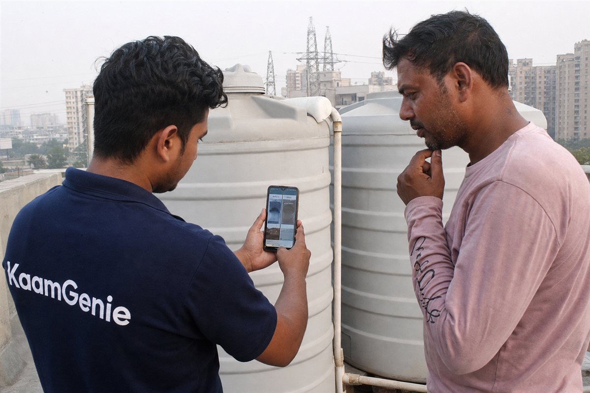 A worker in a navy KaamGenie shirt showing before-and-after tank cleaning photos on a phone to a Noida customer