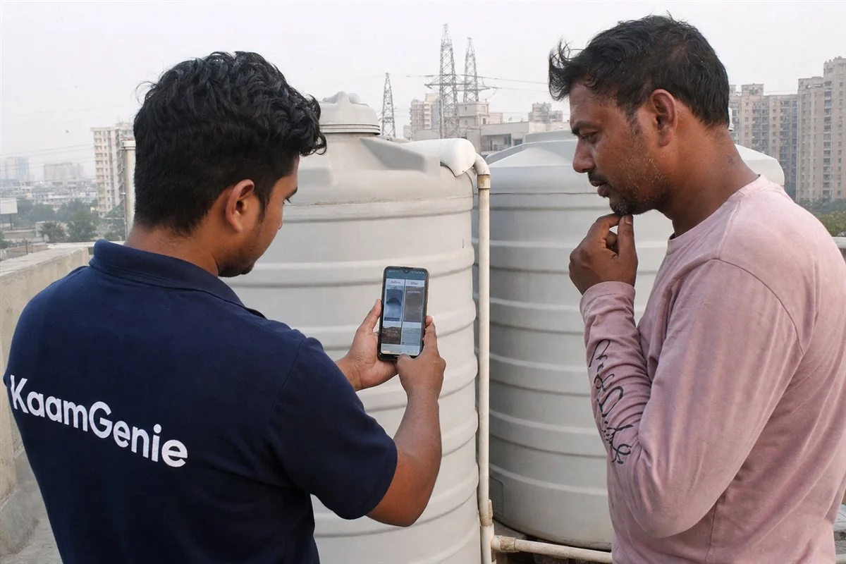 A worker in a navy KaamGenie shirt showing before-and-after tank cleaning photos on a phone to a Noida customer