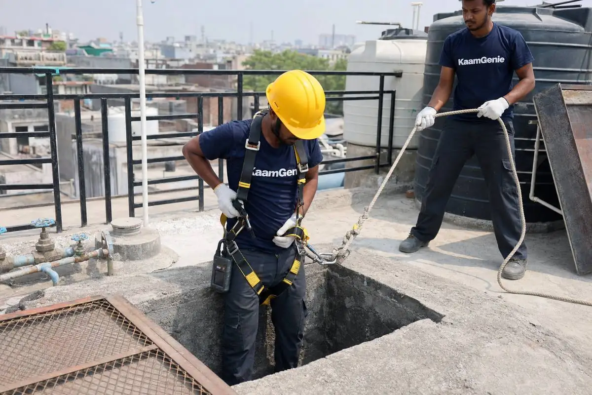 KaamGenie crew member strapping into a safety harness with gas meter and hard hat before descending into a Delhi sump - standby crew member holds the rope outside