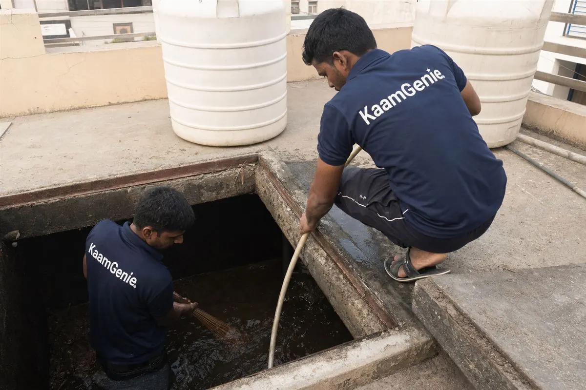 Two KaamGenie workers in navy shirts cleaning a shared underground sump beneath a Delhi builder floor