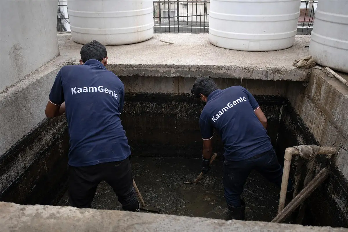 Two KaamGenie crew members in navy shirts cleaning a shared underground sump at the base of a Noida builder-floor building
