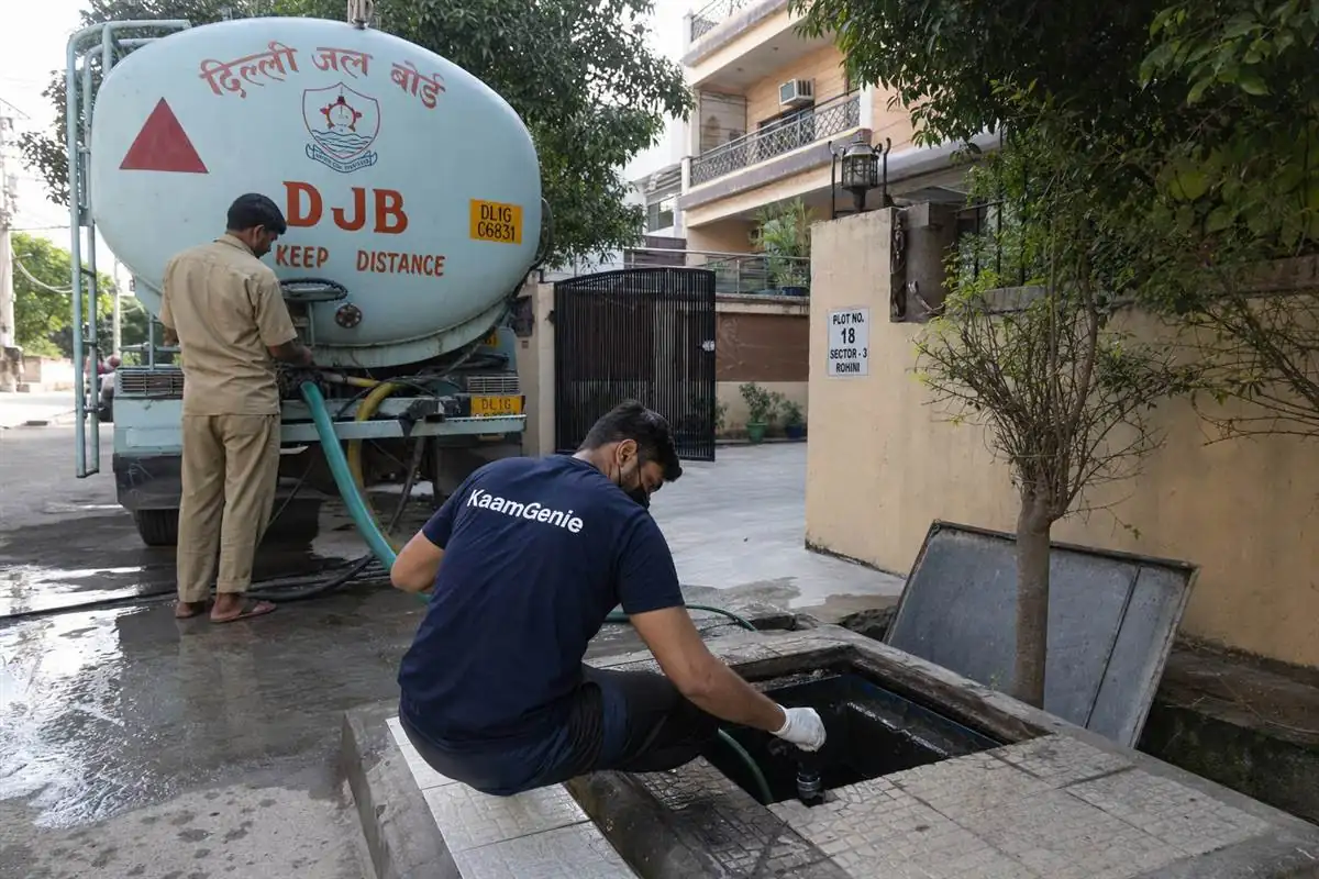 Delhi Jal Board water tanker filling a residential underground sump in a Delhi colony while a KaamGenie technician inspects the tank