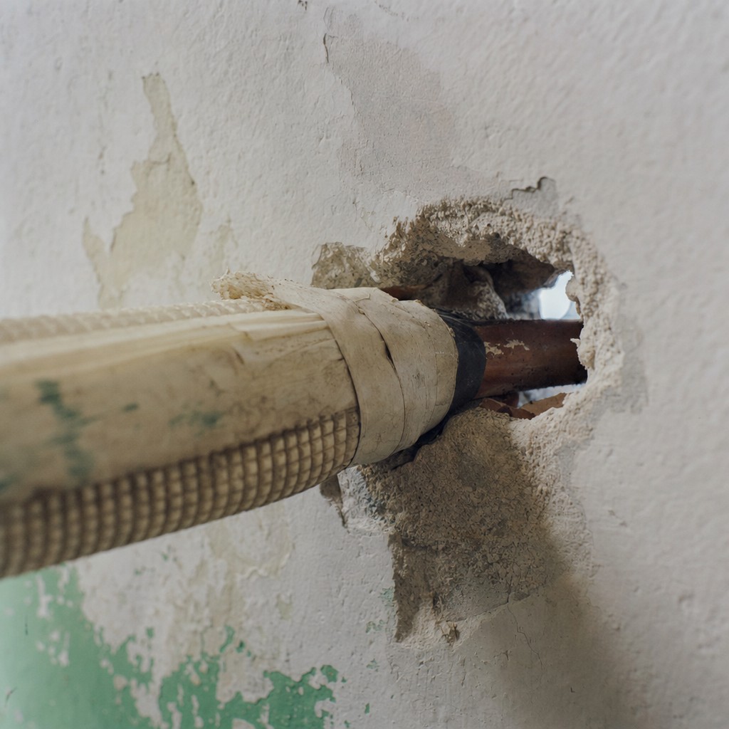 A rat-sized gnaw hole next to a copper AC pipe passing through a Delhi flat wall, with daylight showing through the gap