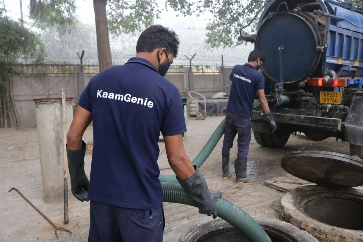 Two KaamGenie workers in navy shirts vacuuming sludge from a large underground reservoir at a Gurgaon society