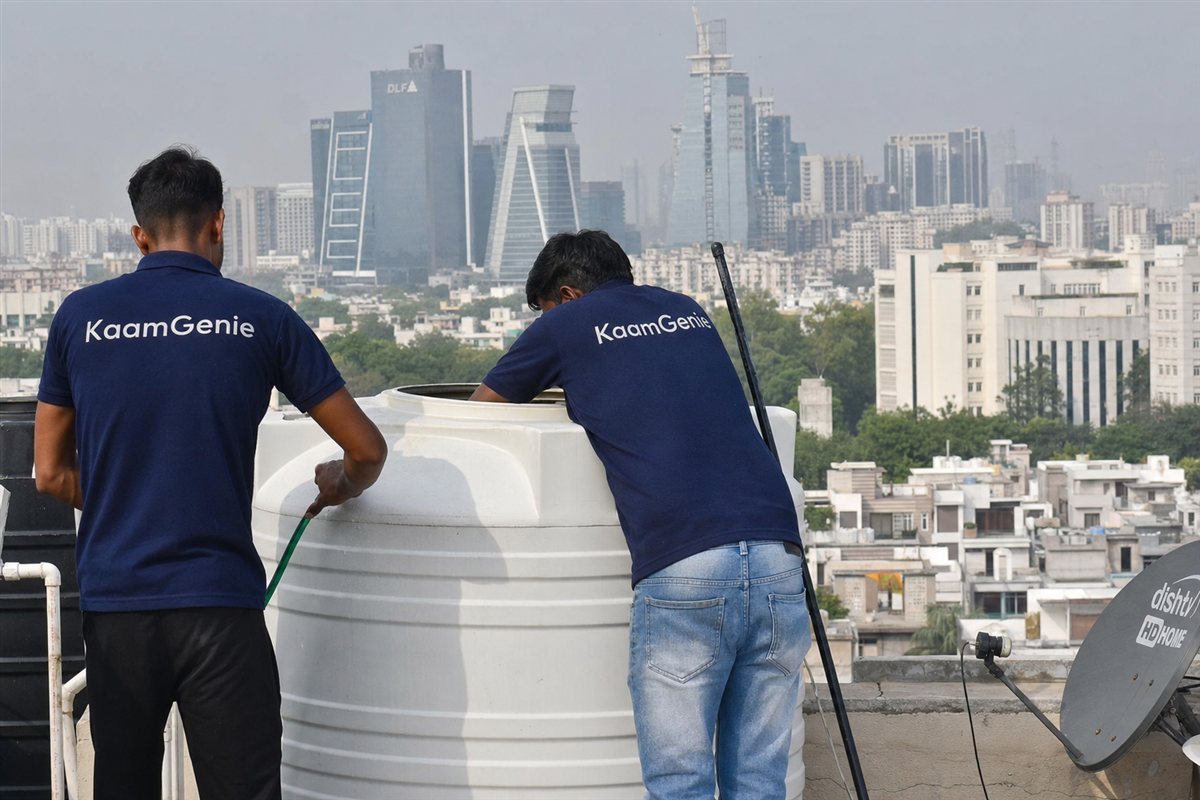 Two KaamGenie workers in navy shirts cleaning a rooftop water tank with the Gurgaon DLF tower skyline behind them