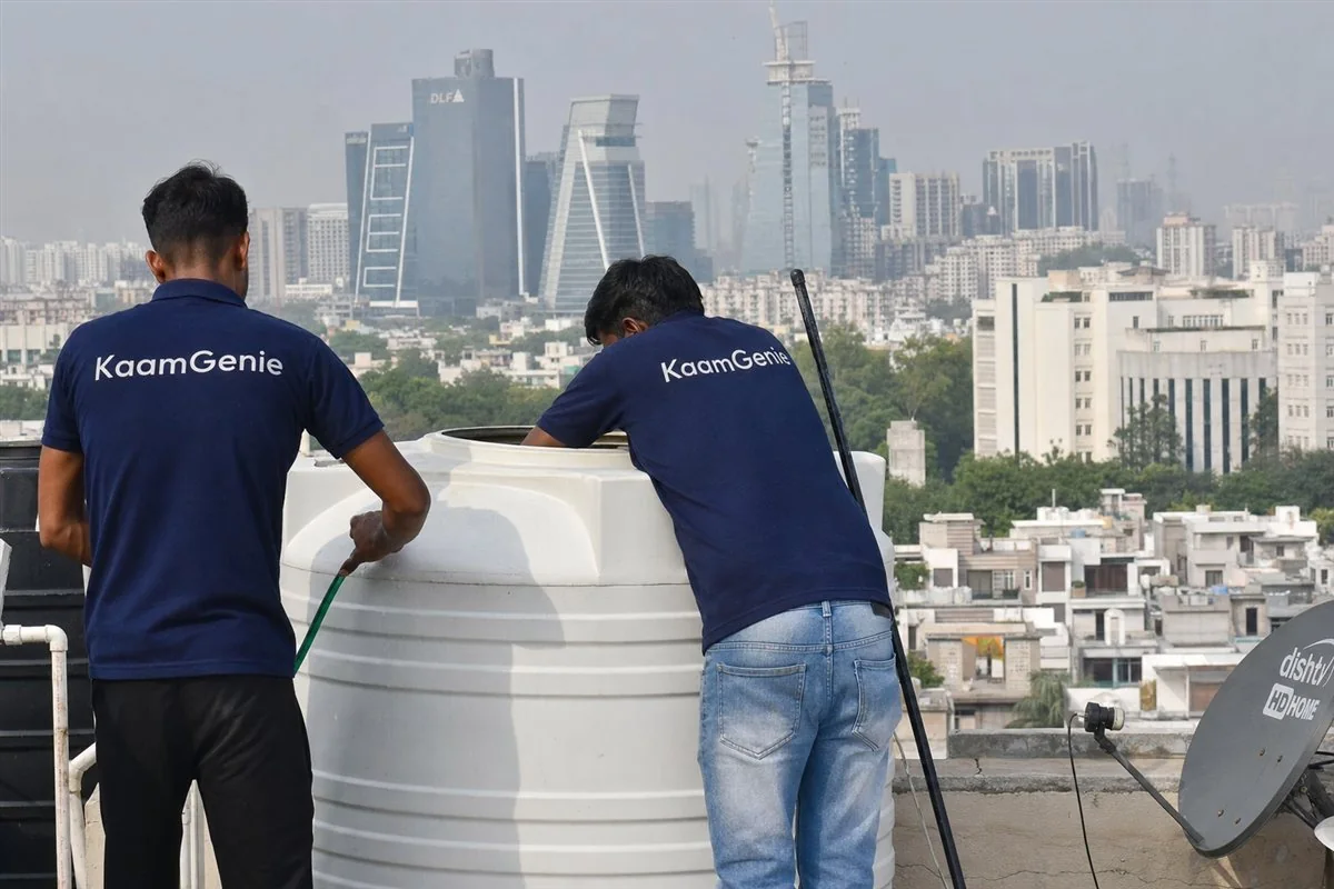 Two KaamGenie workers in navy shirts cleaning a rooftop water tank with the Gurgaon DLF tower skyline behind them