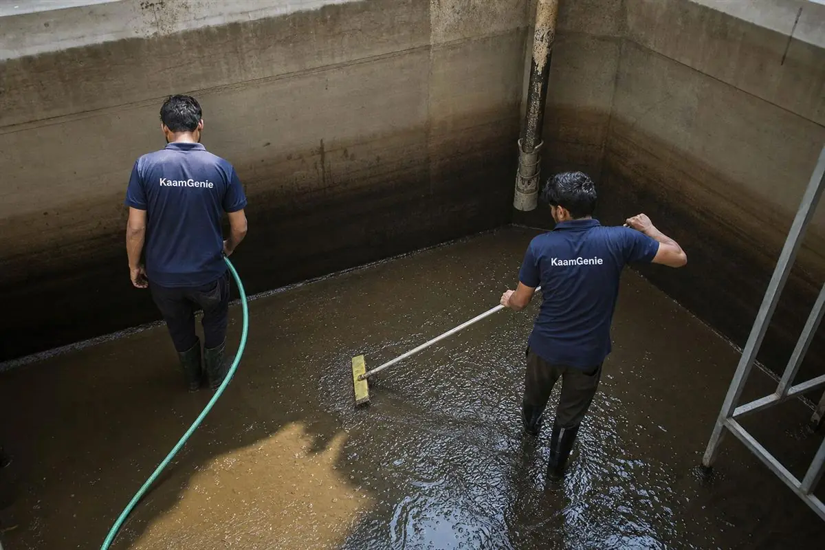 Two KaamGenie workers in navy shirts cleaning a huge underground reservoir at a Dwarka high-rise society, towers visible behind