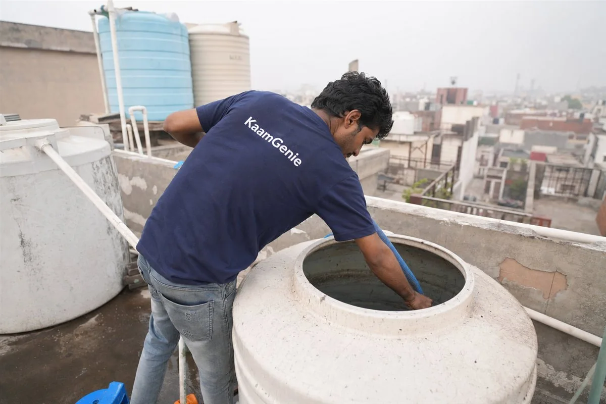 A KaamGenie worker in a navy shirt cleaning a small overhead Sintex tank on the terrace of a plotted Delhi home