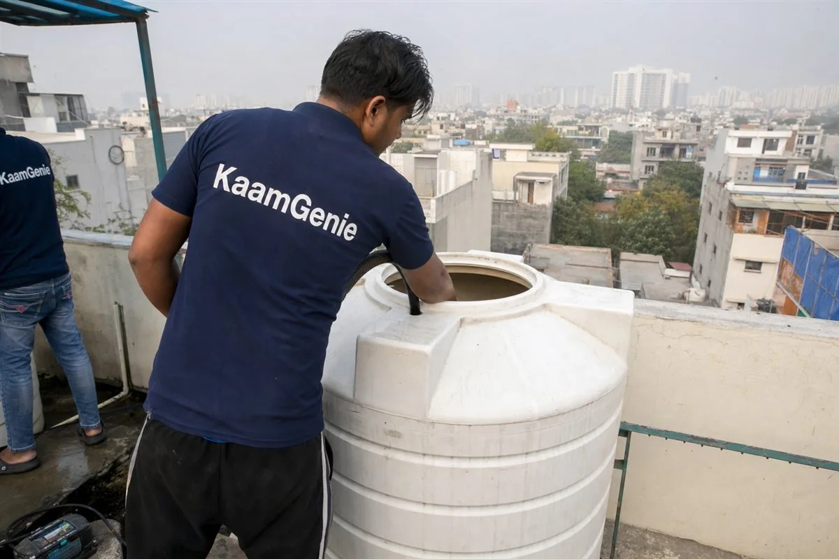 A KaamGenie worker in a navy shirt cleaning a small plastic rooftop tank on a Gurgaon builder-floor terrace
