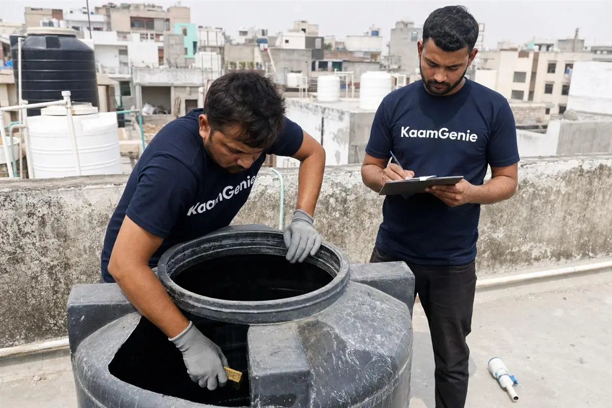 KaamGenie crew member scrubbing the inside of a 1000-litre plastic overhead tank on a Delhi rooftop while a colleague notes the time on a clipboard