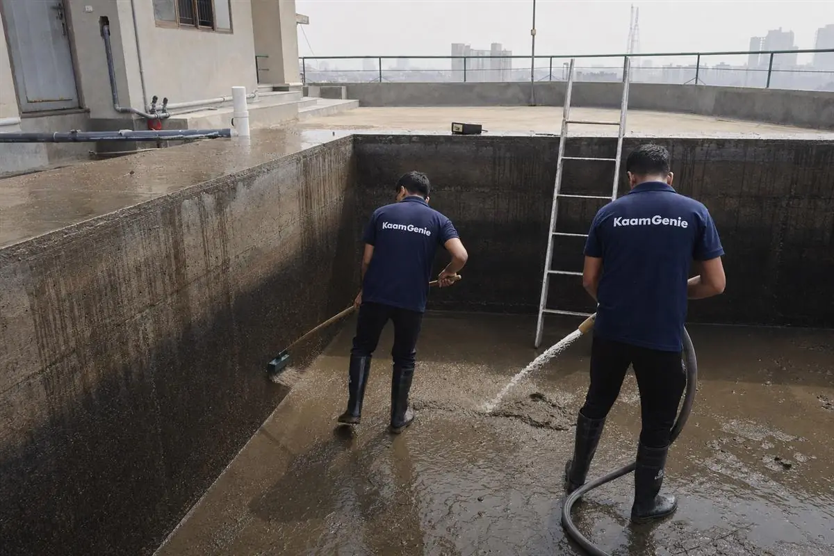 Two KaamGenie workers in navy shirts cleaning a large underground reservoir at a Noida society