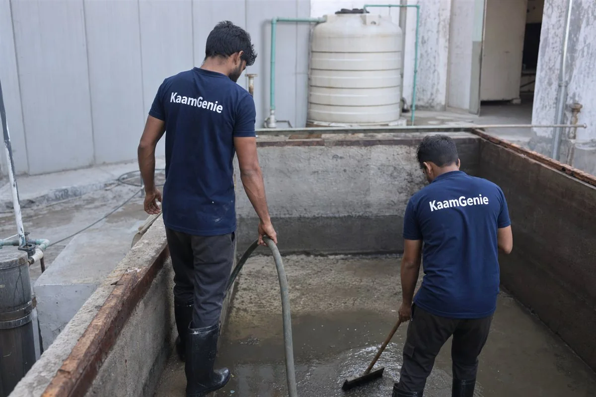Two KaamGenie workers in navy shirts cleaning an underground reservoir at a Gurgaon manufacturing unit with safety harness and gas monitor