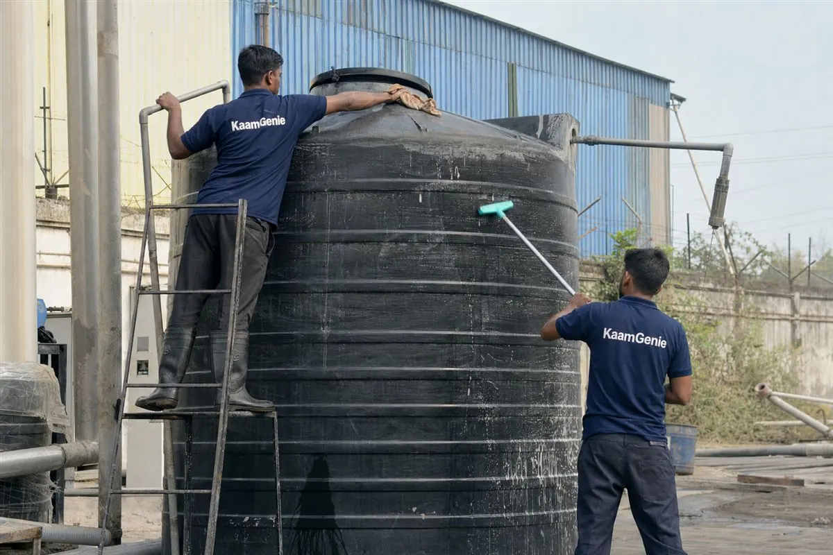KaamGenie crew in navy shirts cleaning a large industrial water storage tank at a Noida factory with sheds behind