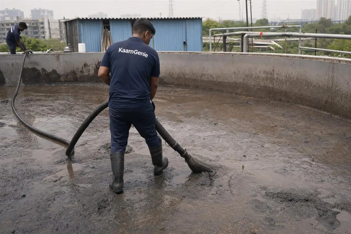 A worker in a navy KaamGenie shirt vacuuming heavy sediment from a drained industrial water tank at a Noida factory