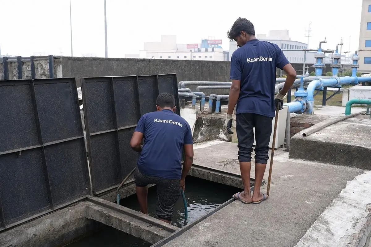 Two workers in navy KaamGenie shirts cleaning a large underground reservoir at a Noida manufacturing unit, one inside and one at the entry as standby