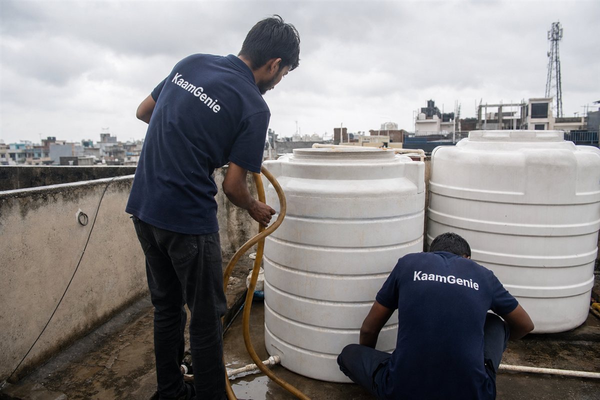 Two KaamGenie crew members in navy shirts cleaning a rooftop water tank during cloudy monsoon weather on a wet Delhi terrace