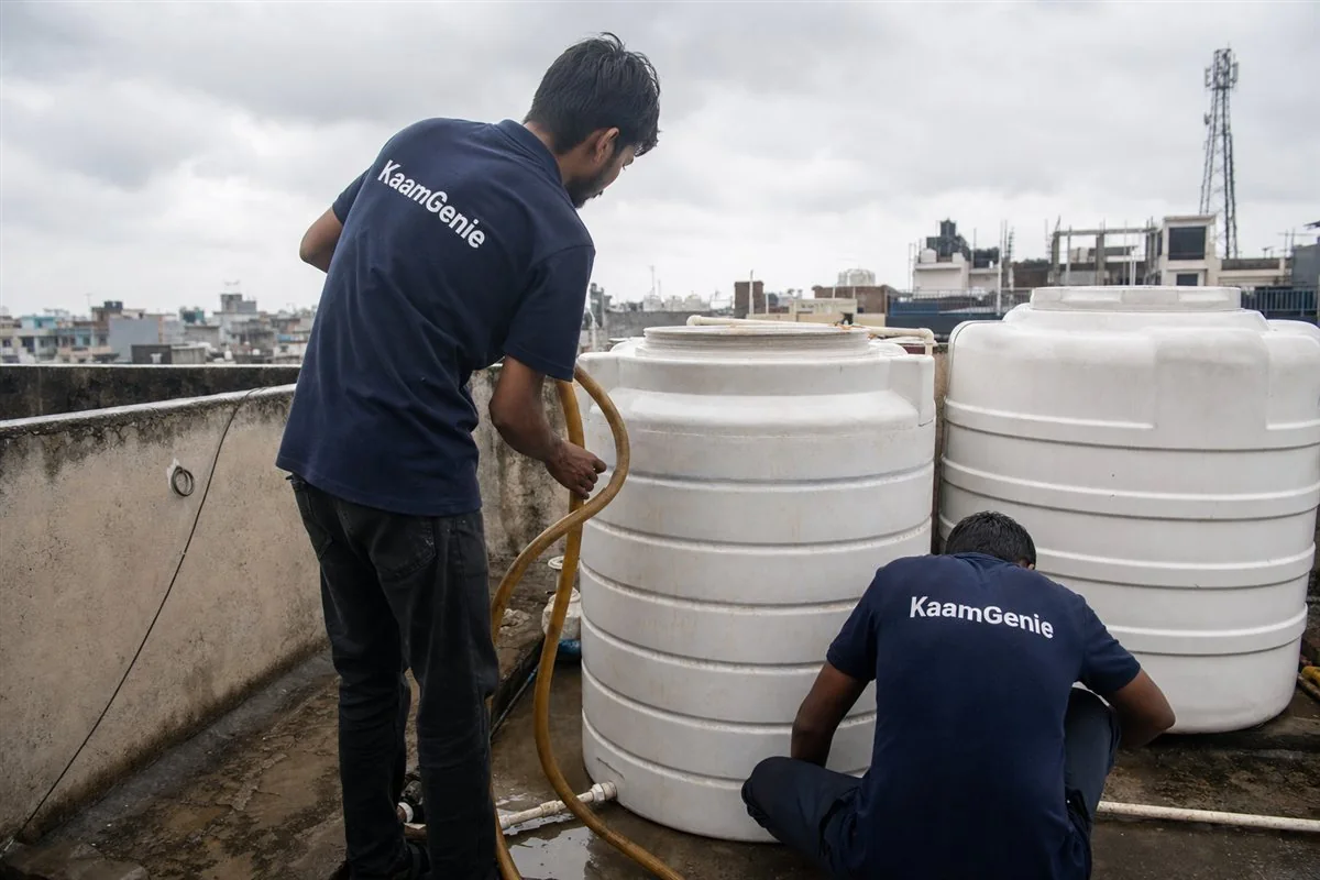 Two KaamGenie crew members in navy shirts cleaning a rooftop water tank during cloudy monsoon weather on a wet Delhi terrace