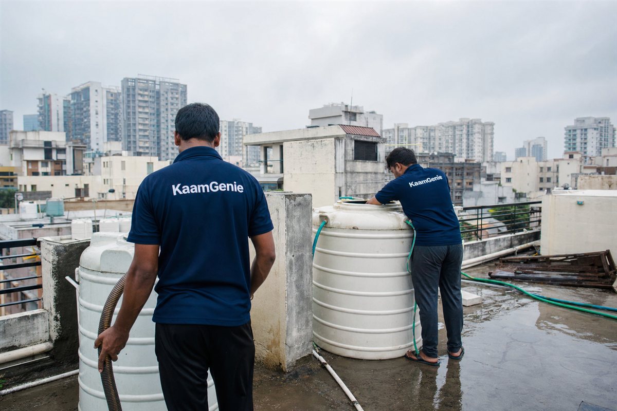 Two KaamGenie workers in navy shirts cleaning a rooftop water tank on a wet Gurgaon terrace under a cloudy monsoon sky, towers in the background