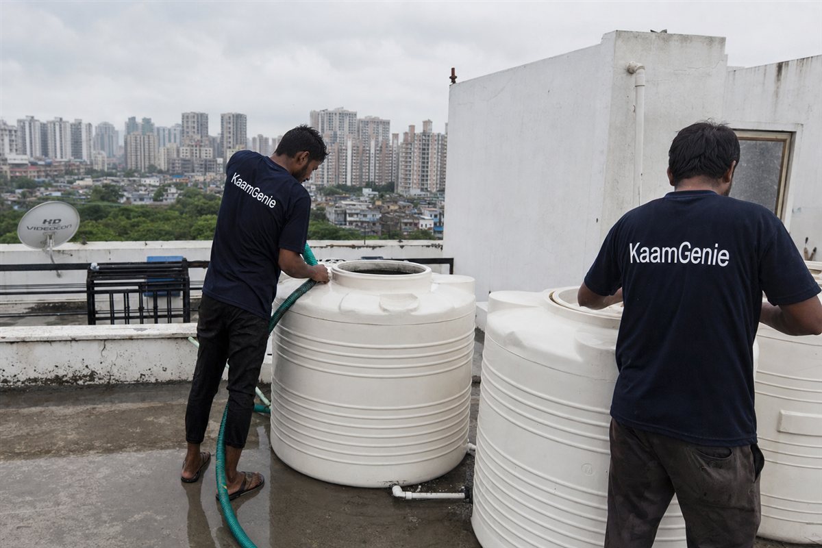 Two KaamGenie workers in navy shirts cleaning a rooftop water tank during cloudy monsoon weather on a wet Noida terrace