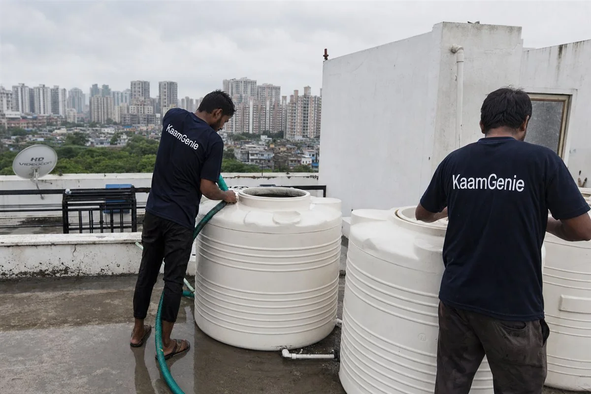 Two KaamGenie workers in navy shirts cleaning a rooftop water tank during cloudy monsoon weather on a wet Noida terrace