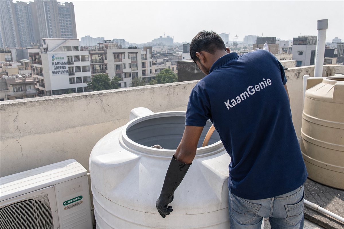 A KaamGenie worker in a navy shirt hand-scrubbing the inside of a builder-floor rooftop drum tank in an old Noida sector