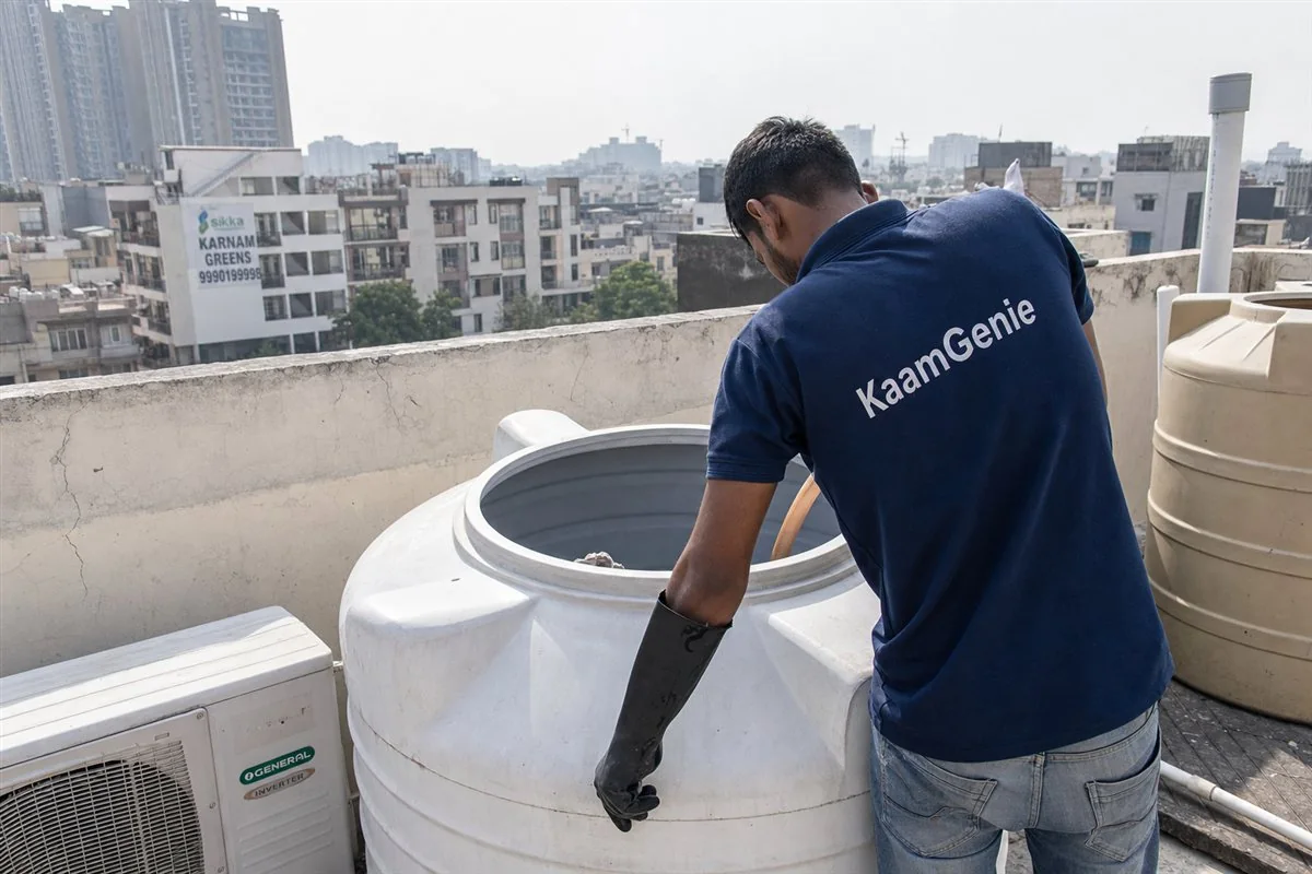 A KaamGenie worker in a navy shirt hand-scrubbing the inside of a builder-floor rooftop drum tank in an old Noida sector