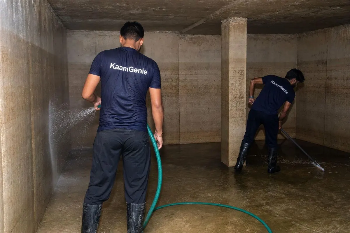 Two KaamGenie workers in navy shirts cleaning a large high-rise underground reservoir in a Noida Extension township