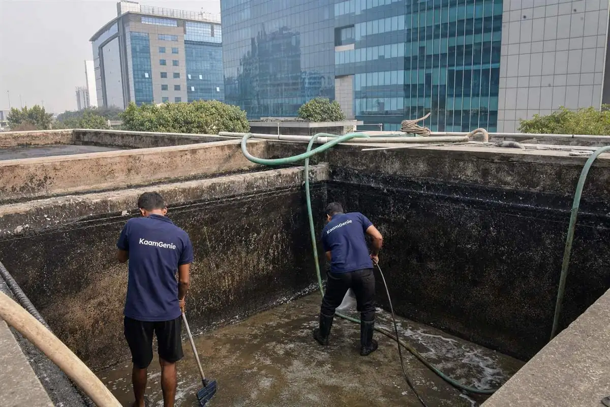 KaamGenie crew in navy shirts cleaning a large underground water reservoir at a Sector 62 Noida IT-park office tower with glass buildings behind
