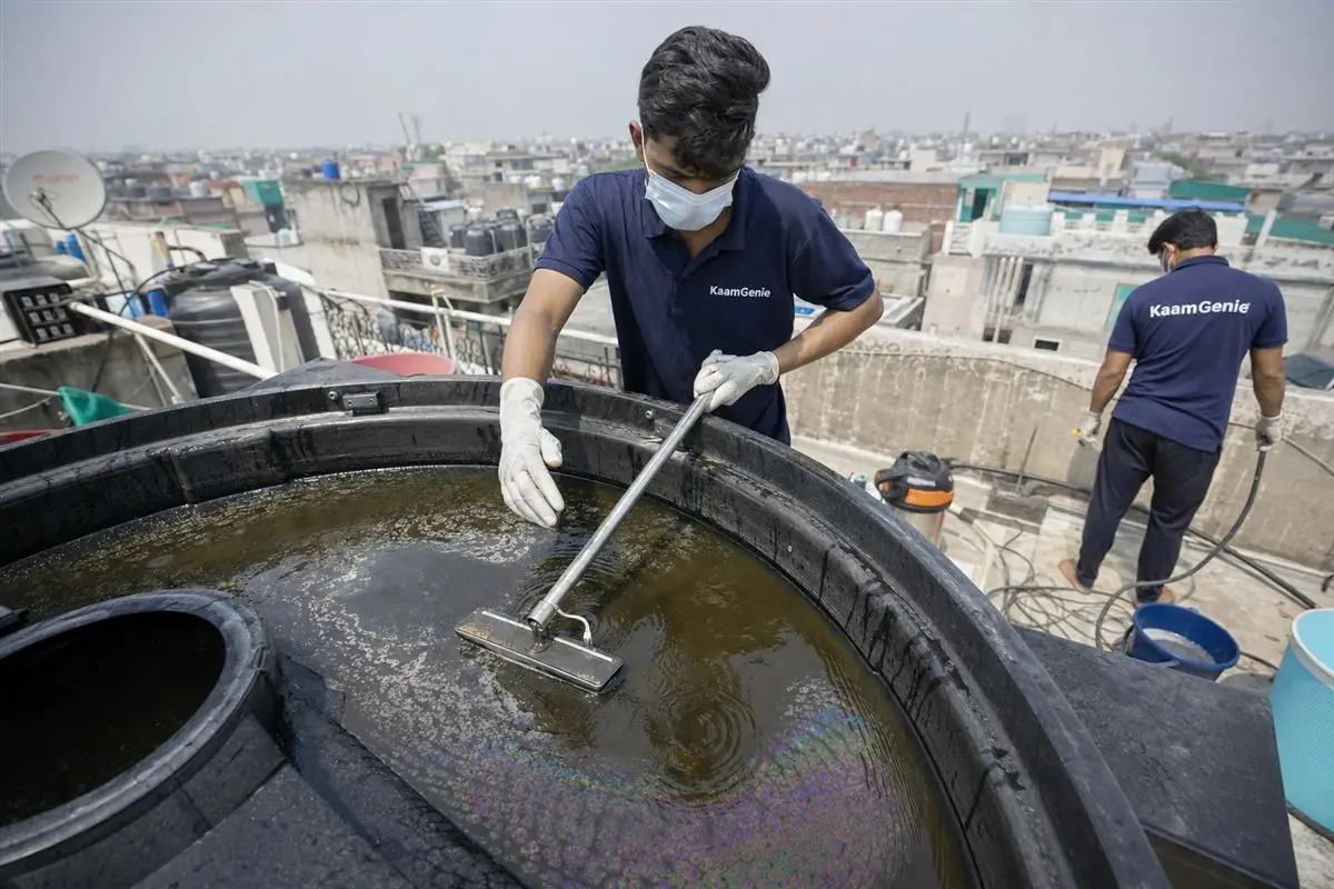 KaamGenie technician skimming an oily rainbow film off the surface of an overhead water tank on a Delhi rooftop