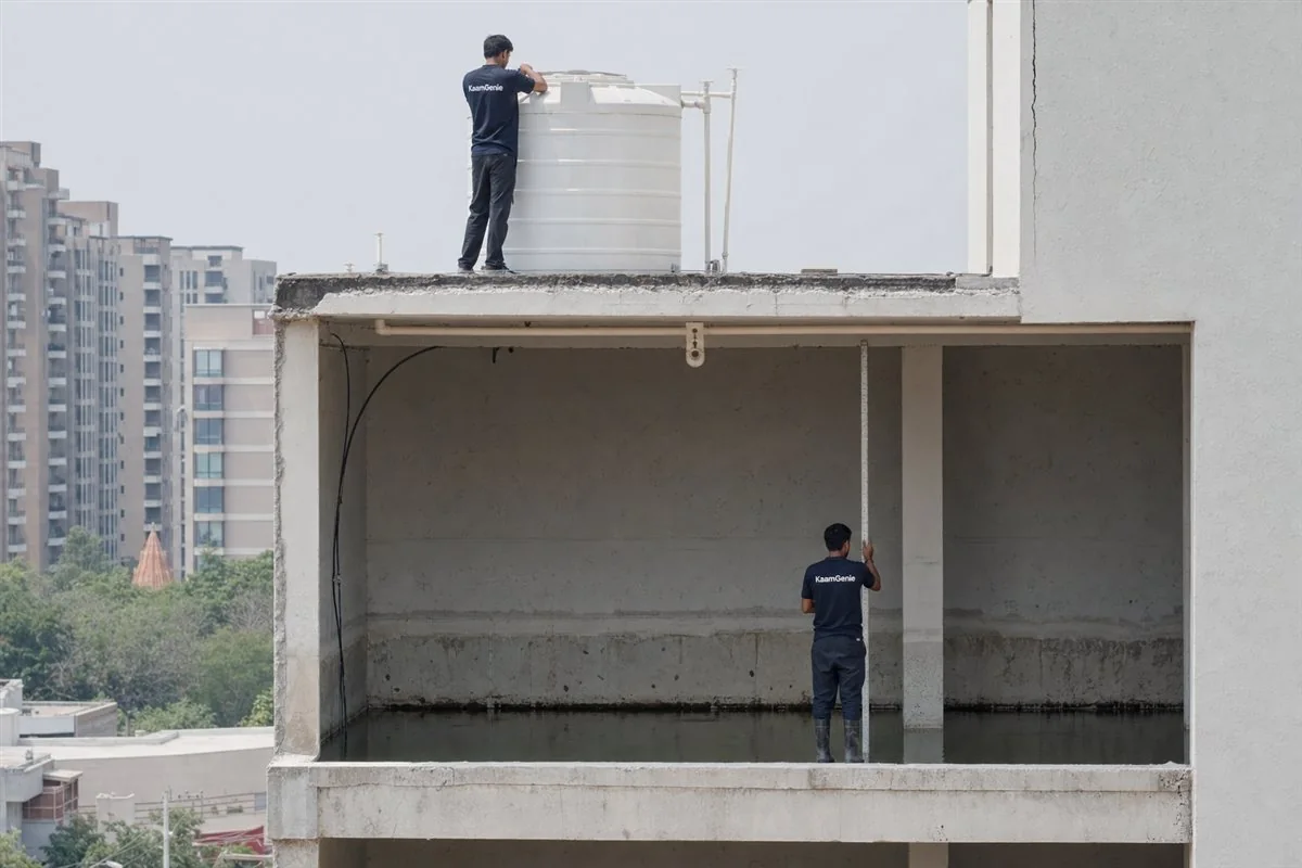 Cross-section of a Gurgaon building showing a rooftop overhead tank and a large underground reservoir, with a KaamGenie worker in a navy shirt at each
