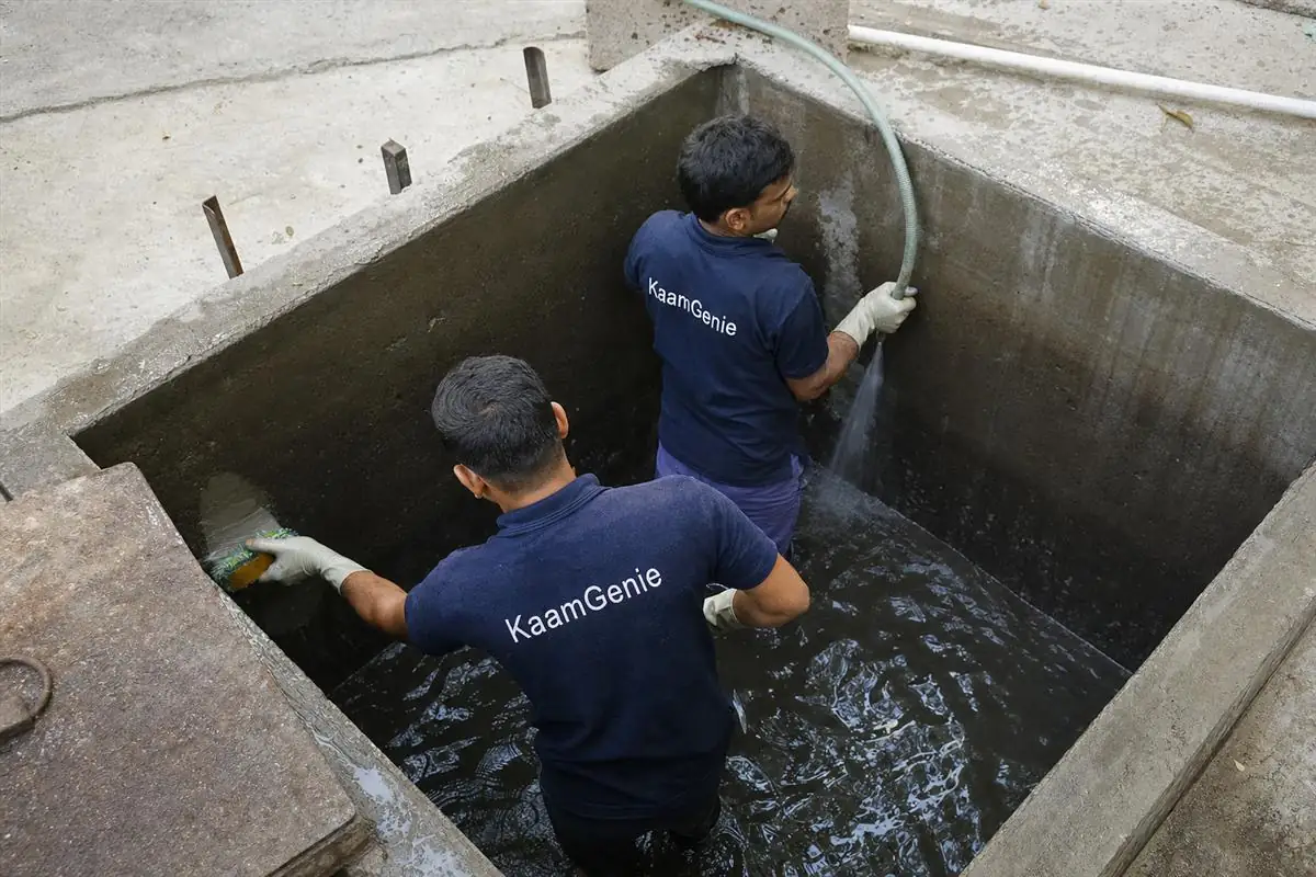 Two KaamGenie workers in navy shirts cleaning a deep underground concrete reservoir at a Gurgaon building, removing settled sediment