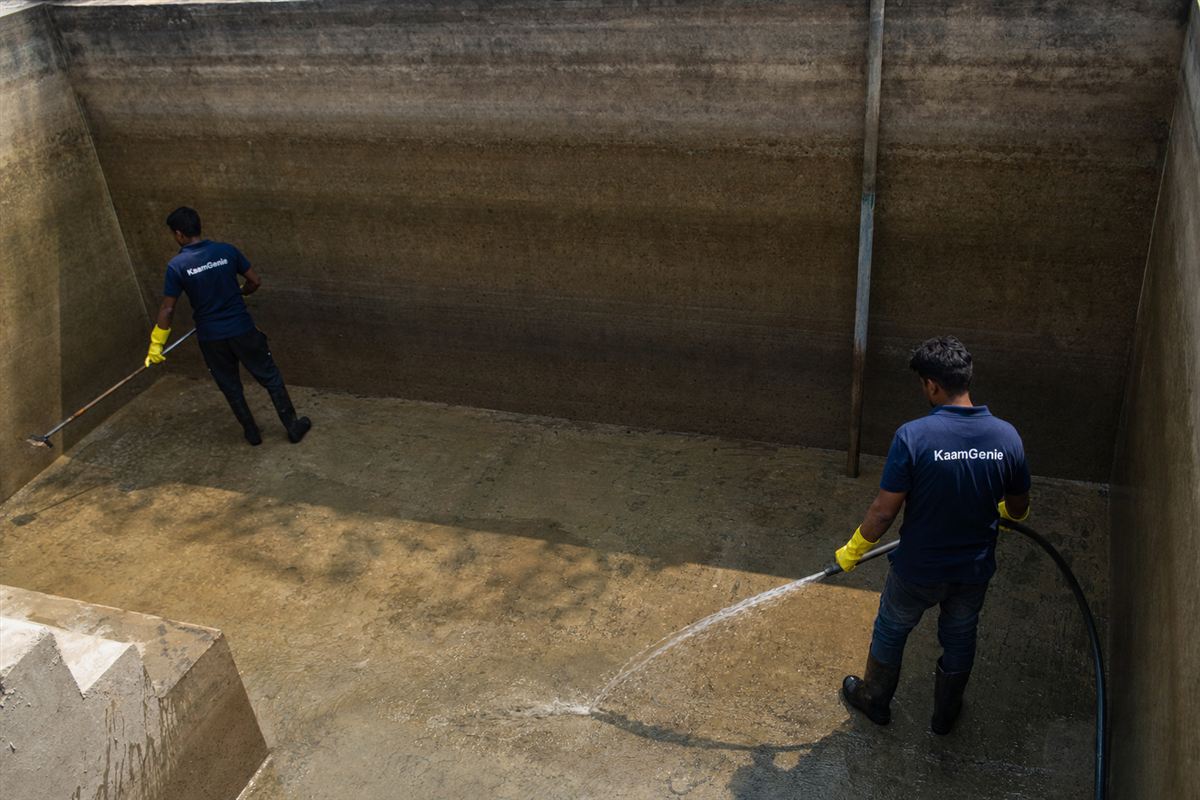 Two KaamGenie workers in navy shirts cleaning a deep underground concrete reservoir at a Noida building, desludging the floor