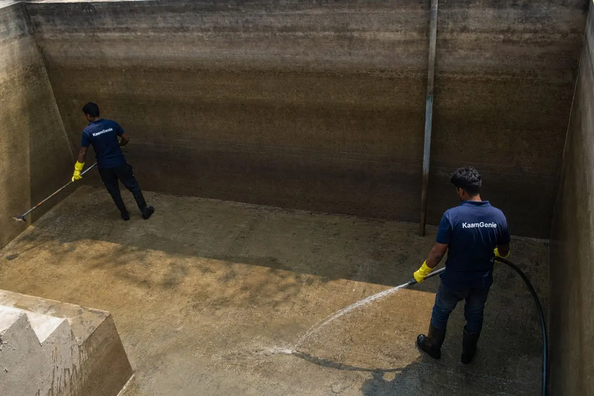 Two KaamGenie workers in navy shirts cleaning a deep underground concrete reservoir at a Noida building, desludging the floor