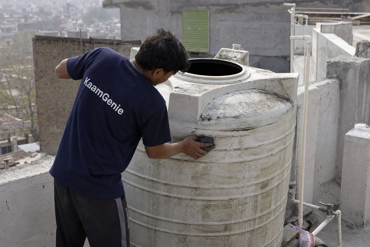 KaamGenie worker in a navy shirt scrubbing a heavily used overhead water tank with thick biofilm on the walls in a Delhi PG