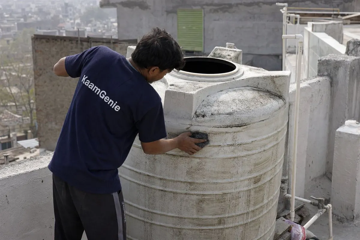 KaamGenie worker in a navy shirt scrubbing a heavily used overhead water tank with thick biofilm on the walls in a Delhi PG