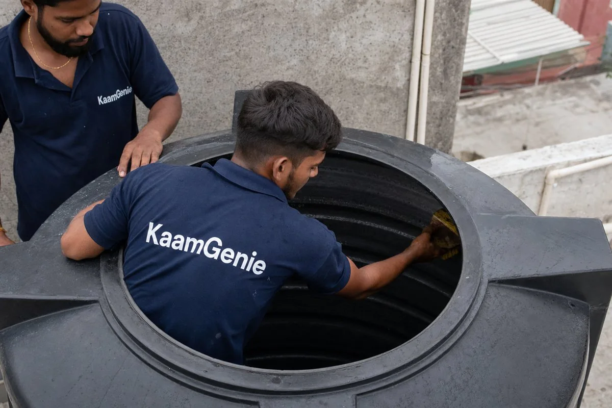 A KaamGenie worker in a navy shirt scrubbing the smooth inside wall of a black plastic Sintex tank on a Gurgaon terrace