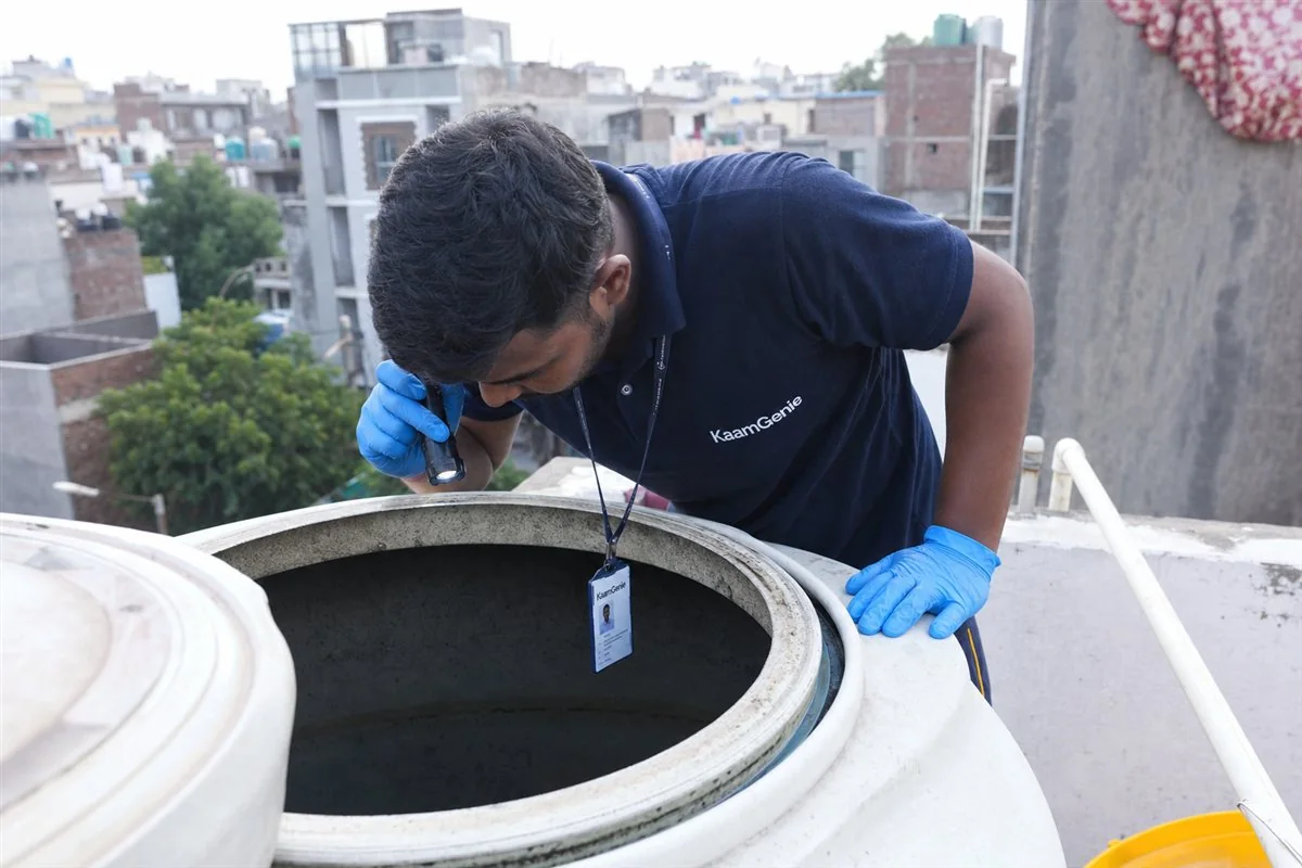 KaamGenie worker in navy uniform inspecting the inside of an open rooftop water tank with a torch on a Delhi terrace