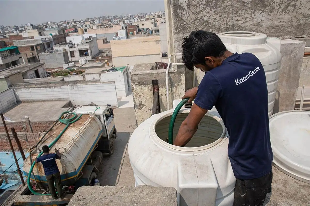 KaamGenie crew member in a navy shirt cleaning a rooftop water tank under harsh Delhi summer sun, a private water tanker visible on the street below