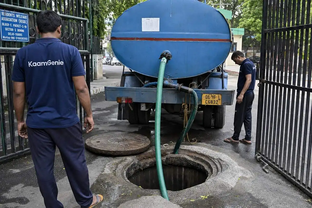 A private water tanker filling a large underground reservoir at a Delhi colony gate while a KaamGenie worker in a navy shirt stands nearby