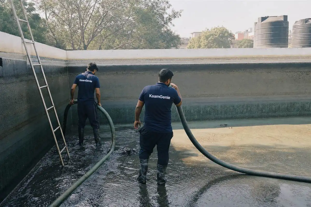 Two KaamGenie workers in navy shirts vacuuming thick sludge from the floor of a large underground reservoir in Delhi