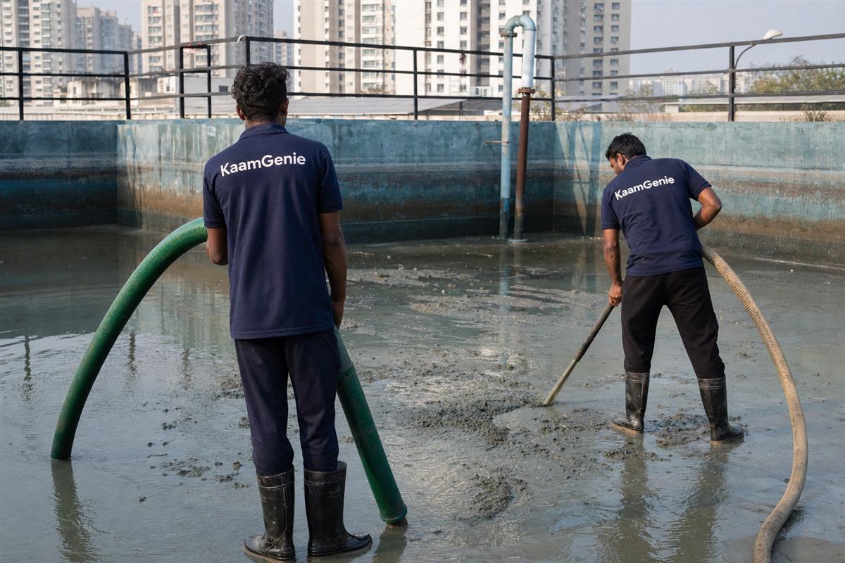 Two KaamGenie workers in navy shirts vacuuming sludge from a society underground reservoir in Noida