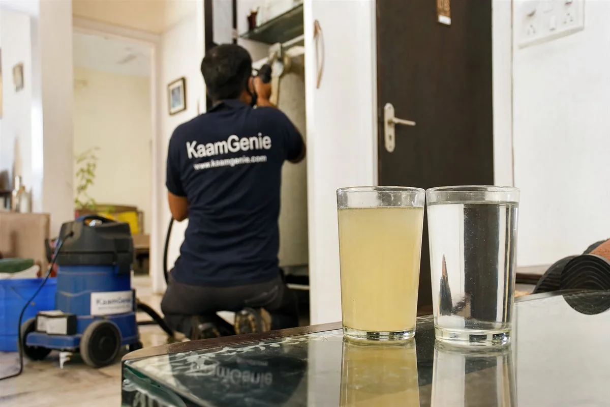 Close-up of yellowish water in a clear glass next to a clean glass, with a KaamGenie technician in navy-blue branded shirt in a Delhi home