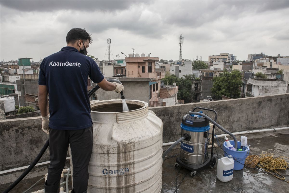KaamGenie technician cleaning a rooftop water tank in early morning Delhi light with monsoon clouds in the background, documentary style