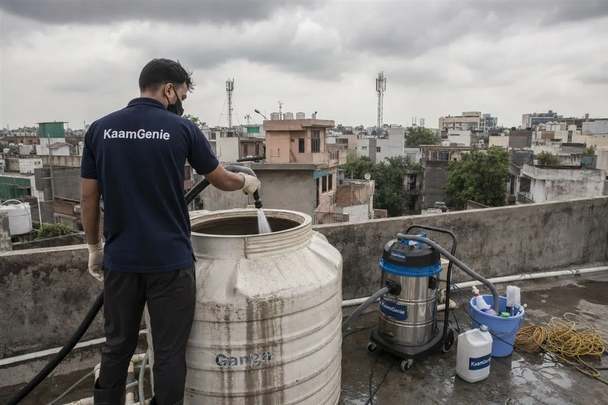 KaamGenie technician cleaning a rooftop water tank in early morning Delhi light with monsoon clouds in the background, documentary style