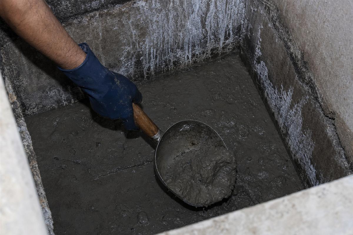 KaamGenie crew member inside an underground reservoir manually scooping settled sludge into a bucket while a colleague stands by at the opening