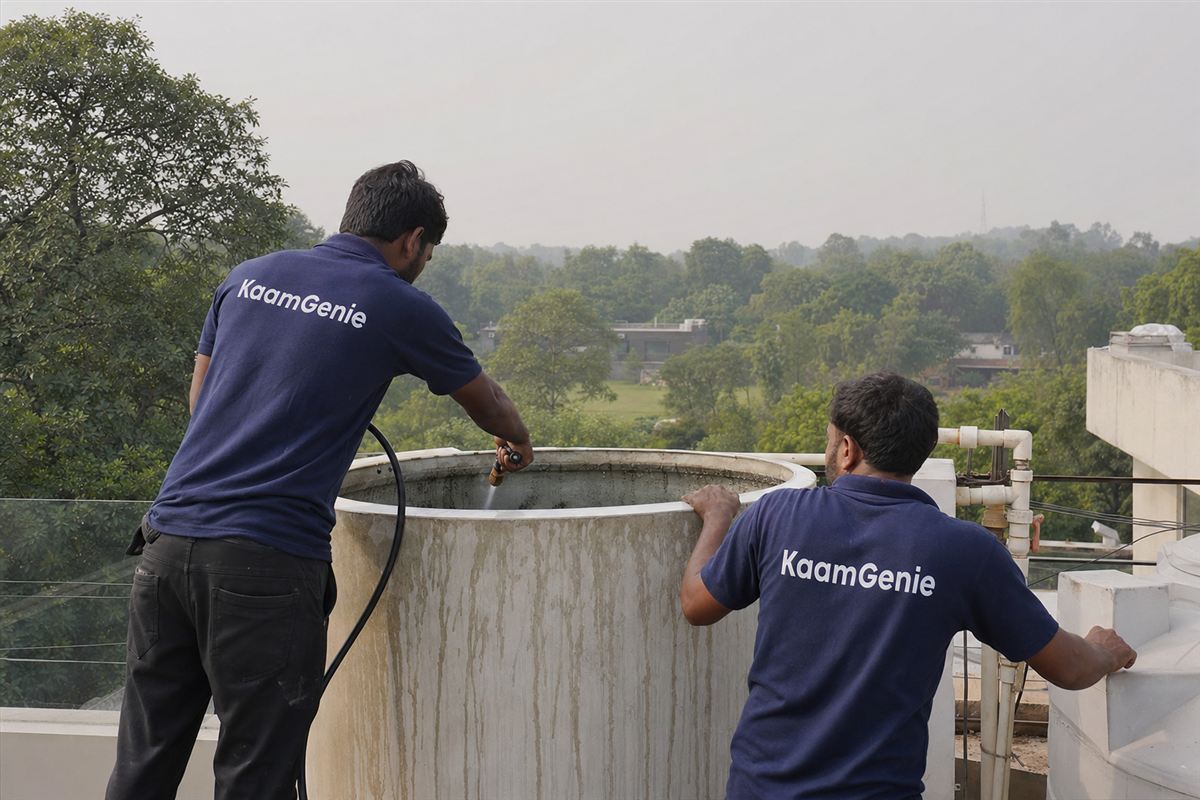 Two KaamGenie crew in navy shirts cleaning a rooftop tank at a leafy South Delhi farmhouse near Chattarpur