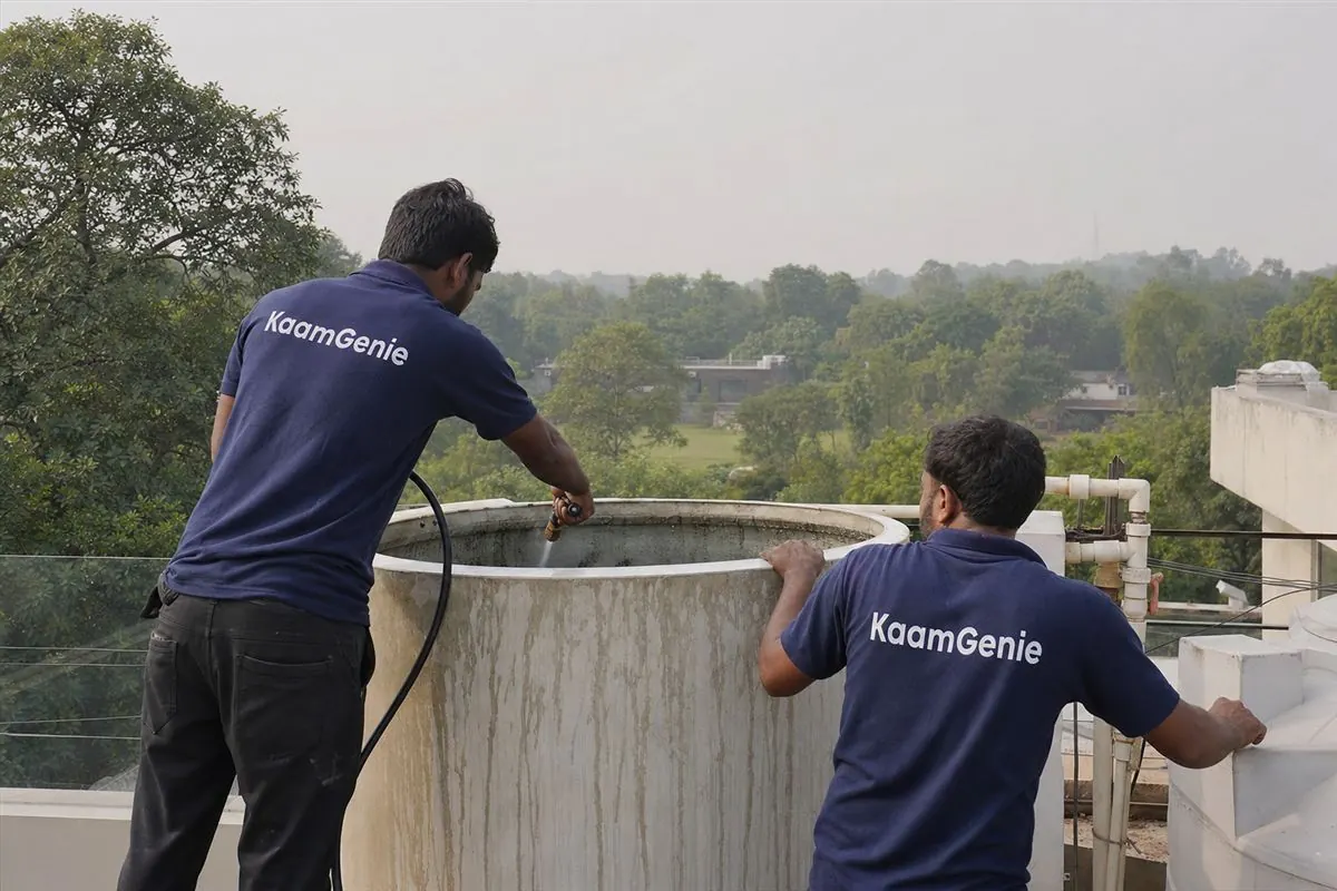 Two KaamGenie crew in navy shirts cleaning a rooftop tank at a leafy South Delhi farmhouse near Chattarpur