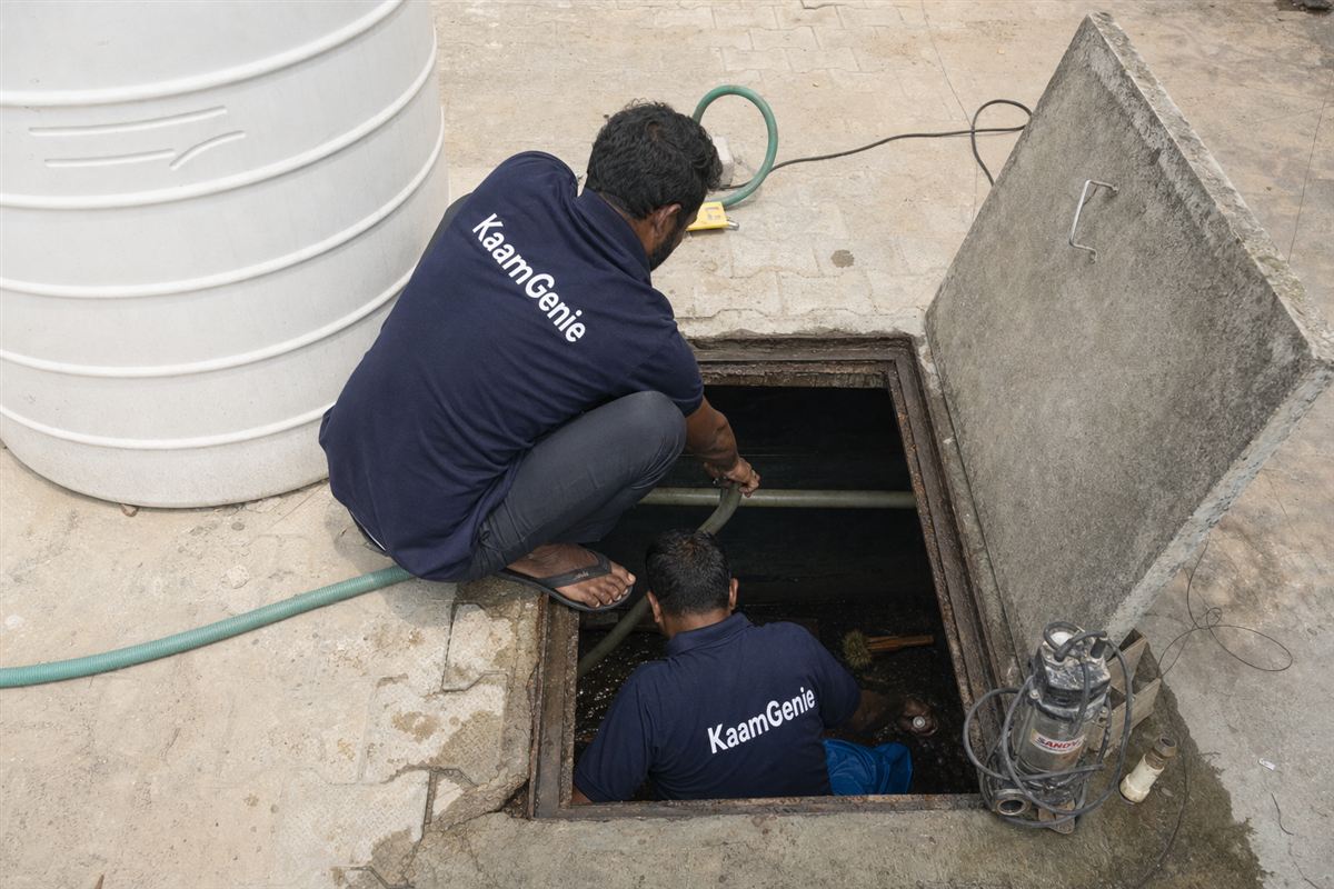 Two KaamGenie crew members in navy shirts cleaning a Gurgaon underground sump with a submersible pump and cleaning equipment around them