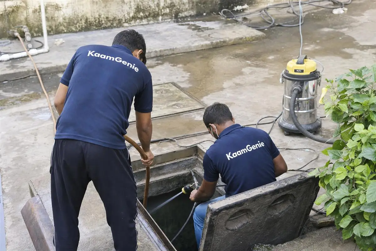 Two KaamGenie crew members in navy shirts cleaning an underground sump at a Noida property, with pump, hose, vacuum and safety equipment laid out around the open reservoir