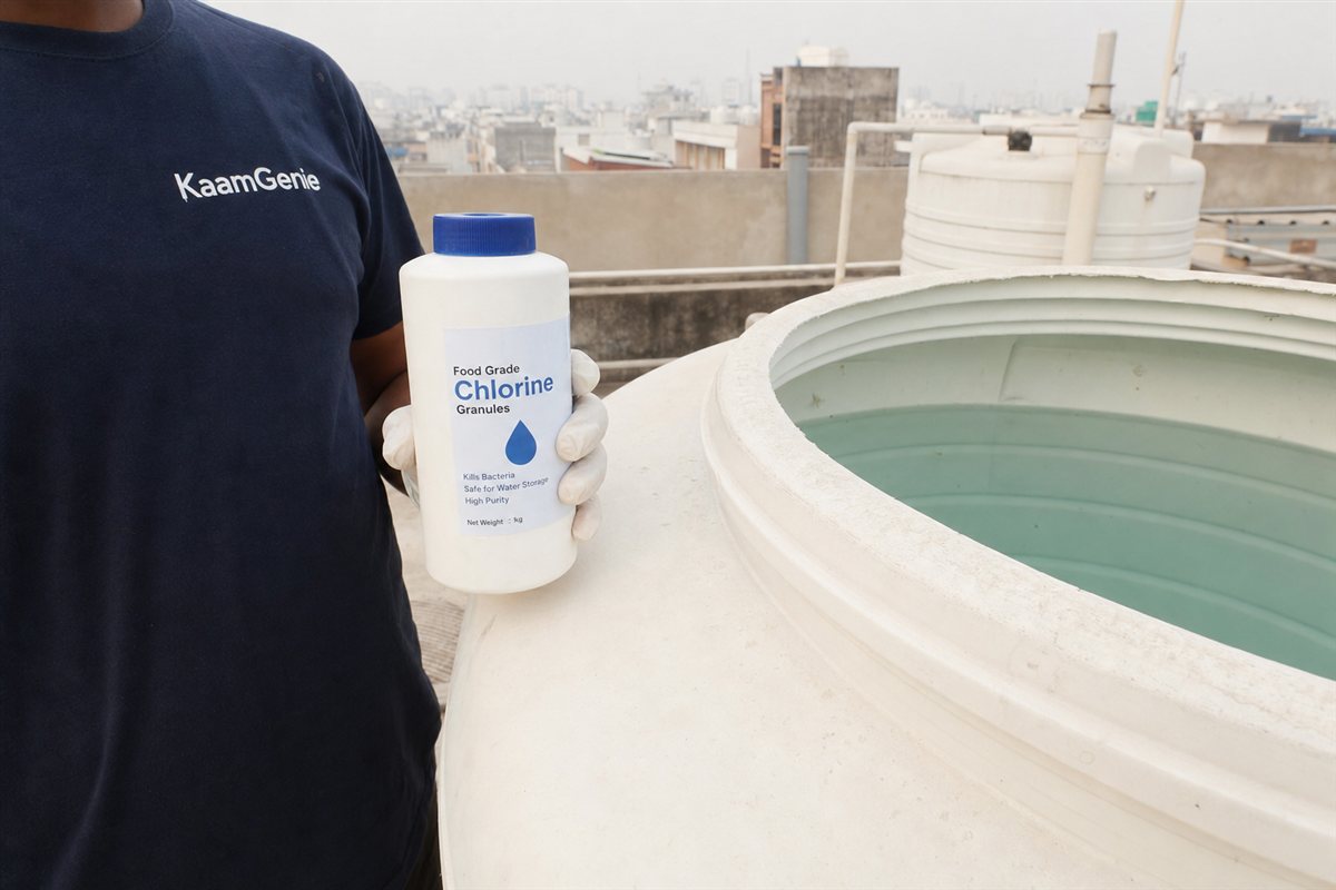 A gloved hand in a navy KaamGenie shirt holding a bottle of food-grade chlorine beside a freshly cleaned Noida water tank